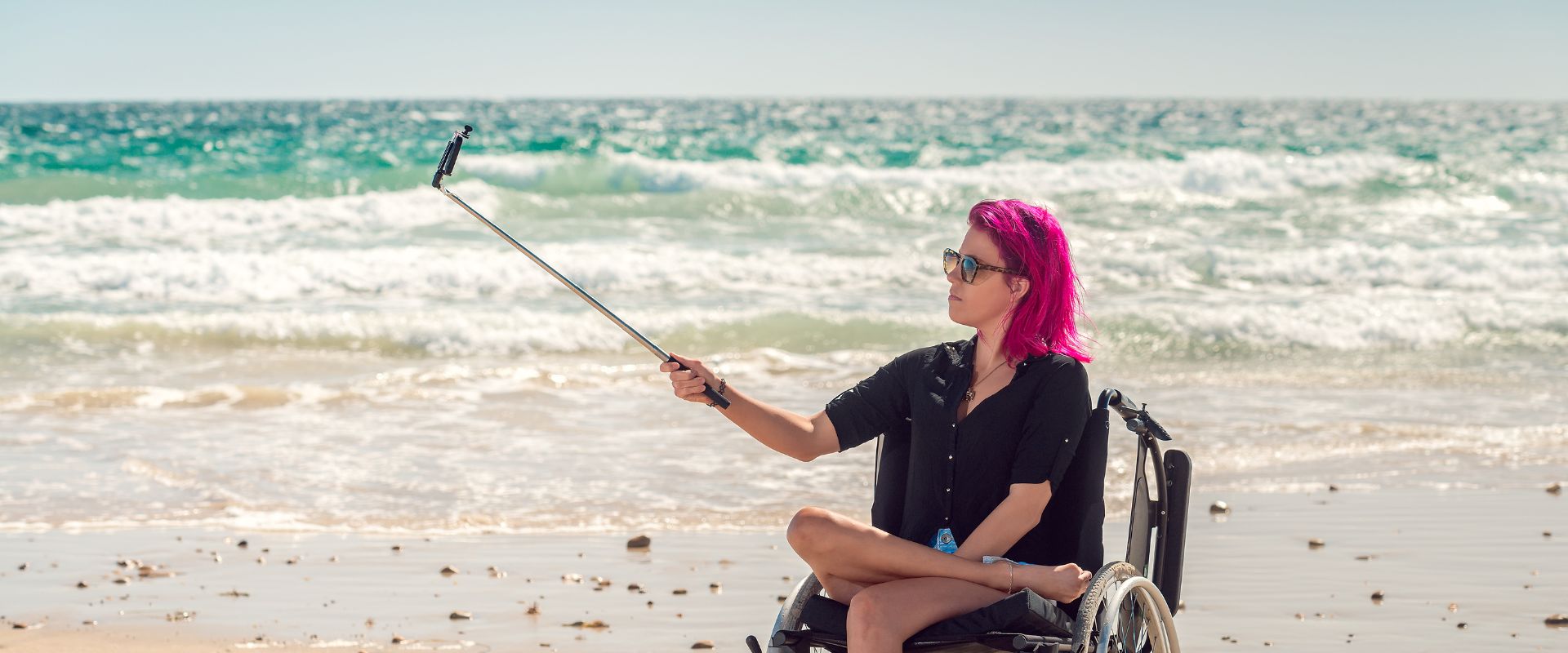 An influencer in a wheelchair at the beach uses a selfie stick to film.